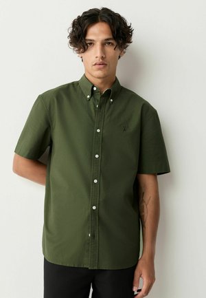 Chemise à manches courtes, verte olive, boutonnée, en coton, avec un col, des boutons sur le devant et un petit emblème sur la poitrine.