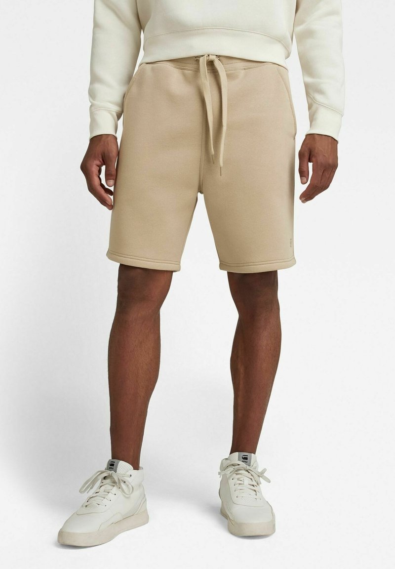 Homme portant un short beige à cordon, un sweat-shirt blanc et des baskets blanches, debout devant un fond blanc uni.