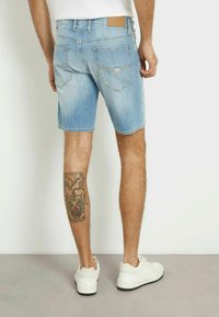 Shorts in denim azzurro chiaro con taglio dritto, dotati di due tasche posteriori e di una patch in pelle tan con il marchio sulla vita. Sneakers bianche visibili.