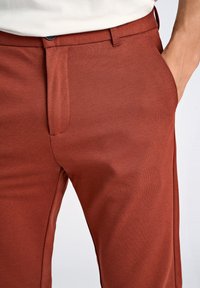 Pantaloni slim-fit marroni realizzati in tessuto testurizzato, dotati di tasche laterali e chiusura con zip. Design pulito con superficie liscia.