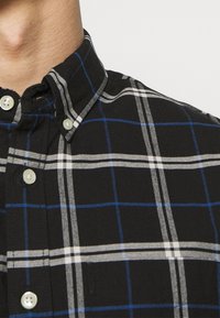 Zwarte geruite blouse met witte en blauwe ruitpatronen. Button-down kraag met lichtgekleurde knopen en een gestructureerde stof.