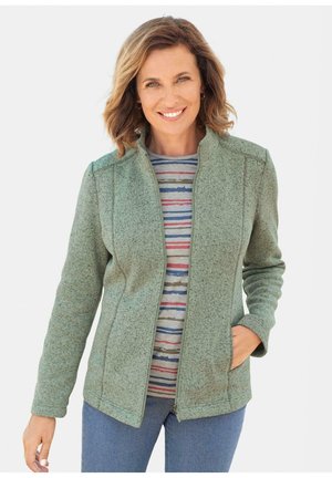 Veste en polaire verte à fermeture éclair avec un col haut, comportant des poches latérales et un tissu texturé. Portée par-dessus une chemise rayée multicolore.