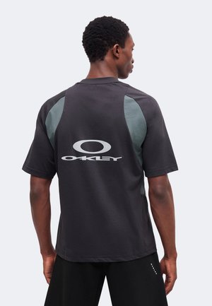 Homme de dos portant un t-shirt Oakley noir avec des panneaux latéraux gris et le logo Oakley imprimé dans le dos.