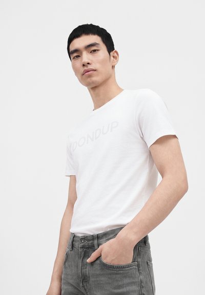 Jeune homme aux cheveux courts noirs portant un t-shirt blanc et un jean gris, debout avec une main dans la poche devant un fond uni.