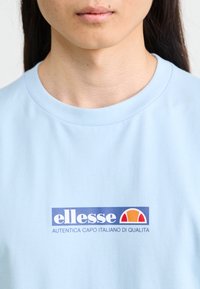 Light blue cotton t-shirt with short sleeves, featuring a large printed logo in blue, orange, and white at the chest.