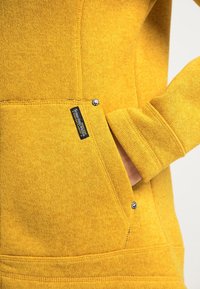 Gelbe Kapuzenjacke aus strukturiertem Strick, mit einer Fronttasche, runden Hardware-Details und einem Etikett, das an der naht der Tasche angenäht ist.