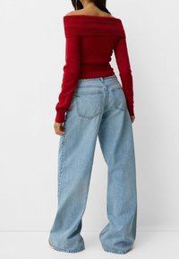 Femme portant un pull rouge à épaules dénudées et un jean à jambes larges bleu clair, debout contre un fond blanc, vue de dos.