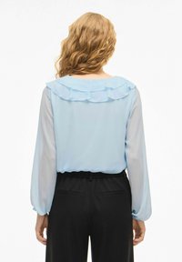 Lyseblå bluse med lange ermer, med en rysjekant rundt halsen. Brukes med svarte bukser med elastisk midje. Glatt stofftekstur.