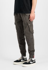 Calças cargo verde-oliveira escuras com vários bolsos, um corte ajustado e punhos elásticos, combinadas com sapatilhas pretas com detalhes brancos.