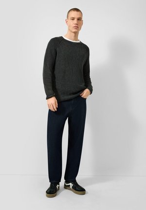 Jonge man staand met een donkergrijze gestructureerde trui, wit onderhemd, marineblauwe broek en zwarte sneakers met witte strepen op een witte achtergrond.
