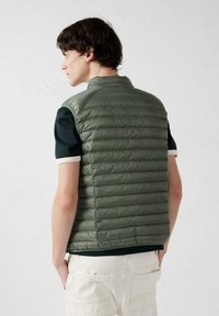 Groene puffer bodywarmer met horizontaal doorgestikt patroon, lichtgewicht materiaal, opstaande kraag en een aansluitend ontwerp over een donker shirt.