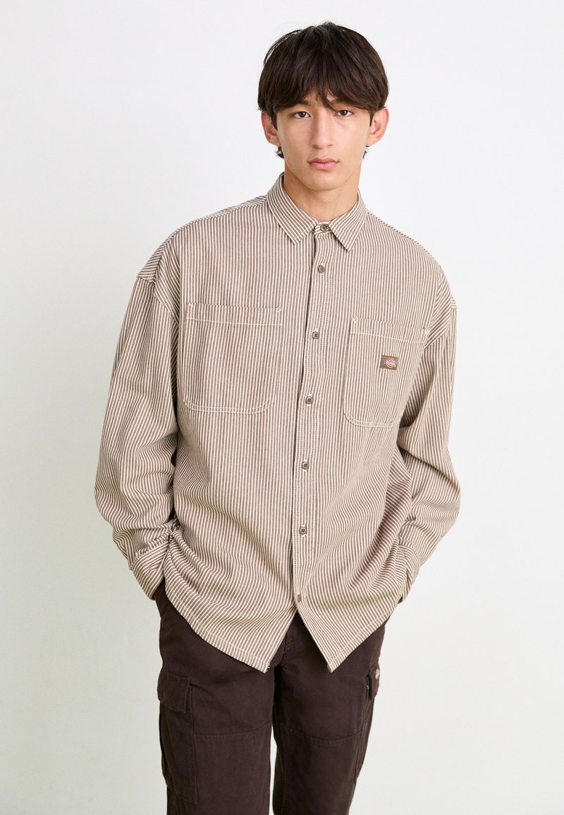 Brown and cream striped long-sleeve shirt with button front, two chest pockets, and a subtle logo patch. Soft fabric and relaxed fit.