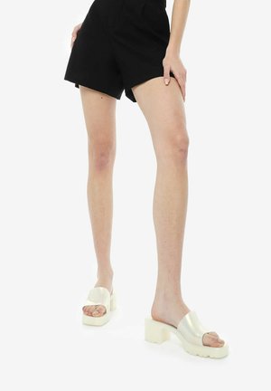 Sandali platform bianchi con finitura lucida, cinturino largo sulla punta e tacco spesso, abbinati a pantaloni corti neri, esposti su uno sfondo semplice.