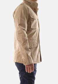 Giacca in canvas beige con colletto alto, diverse tasche e toppe sui gomiti; chiusure a bottoni e zip; design strutturato e vestibilità casual.