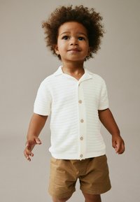 Vit texturerad kortärmad skjorta med krage, fem ljusa knappar, kombinerad med ljusbruna shorts. Barn står med armarna något utsträckta.