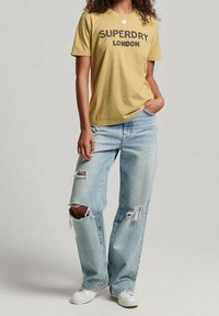 Kvinna som bär en senapsgul Superdry London T-shirt, ljusblå ripped jeans, vita sneakers, stående med en hand i fickan.