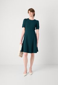 Short-sleeved teal dress with a fitted bodice and flared skirt, featuring decorative buttons on sleeves. Model holds a cream handbag.