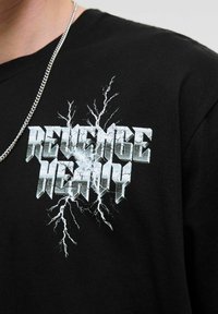 Black cotton t-shirt featuring a cracked metallic graphic with the text "REVENGE HEAVY," accented by lightning-like designs around it.