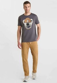 Camiseta gris con un gráfico de un oso con un sombrero beige, combinada con pantalones cargo beige. El conjunto se complementa con zapatillas de color claro.