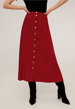 Femme portant une jupe midi rouge avec des boutons argentés sur le devant, associée à des bottes à talons noires et un top noir, debout les mains sur les hanches.