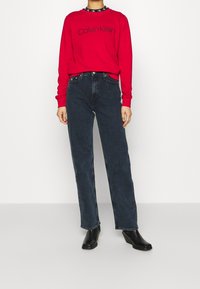 Czerwona bluza z kapturem z logo "Calvin Klein", czarny kołnierz z akcentem, zestawiona z ciemnoniebieskimi prostymi dżinsami i czarnymi botkami na obcasie.