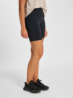 Person iført sorte højtaljede tætsiddende shorts, beige top og sorte athletic sko, stående sidelæns mod en ensfarvet hvid baggrund.