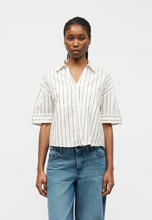 Γυναίκα με πλεξούδες που φοράει λευκό πουκάμισο με μαύρες κάθετες ρίγες και μπλε τζιν, στέκεται μπροστά σε μια απλή λευκή επιφάνεια.