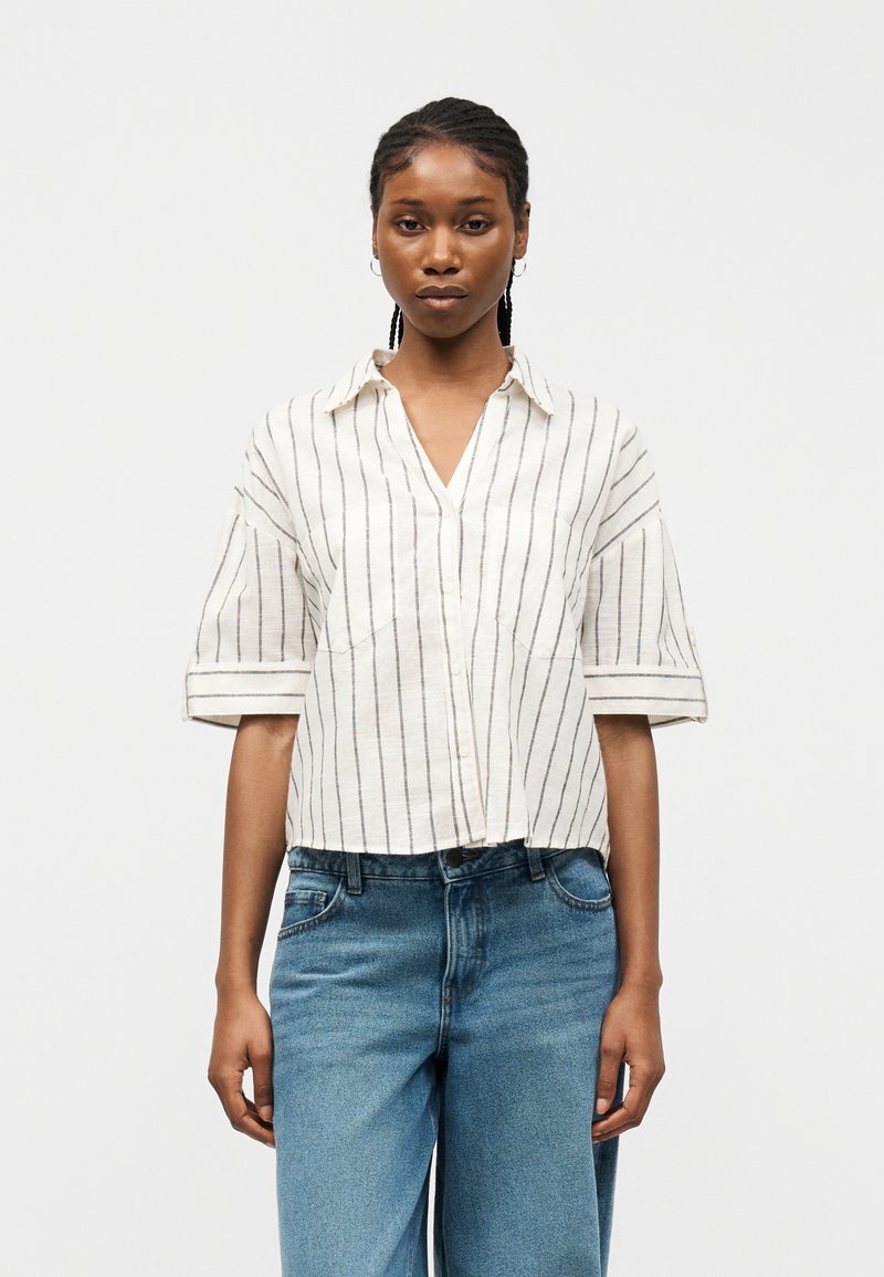 Γυναίκα με πλεξούδες που φοράει λευκό πουκάμισο με μαύρες κάθετες ρίγες και μπλε τζιν, στέκεται μπροστά σε μια απλή λευκή επιφάνεια.