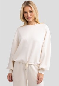 Sudadera color crema con corte holgado, cuello redondo y mangas globo. Presenta un cordón en el dobladillo para un ajuste ajustable.