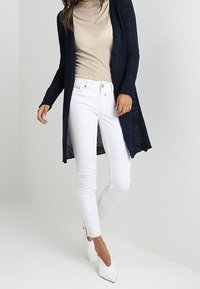 Lång marinblå cardigan med öppenstickad design, kombinerad med vita tajta jeans och spetsiga vita klackar. Slät textur och avslappnad passform.