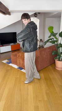 Giacca nera con cappuccio in pelle, pantaloni larghi a righe grigio chiaro e scarpe argento lucide. Ambientazione interna con pavimento in legno e divano marrone.