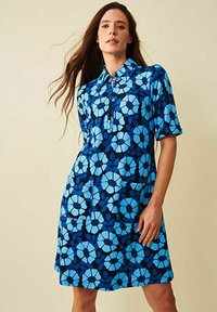 Blommig blå klänning med korta ärmar, krage och fickor. Mönstret består av stora blå blommor på en mörkblå bakgrund.