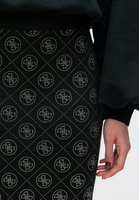 Schwarzer Rock mit einem Rautenmuster, verziert mit metallischen Akzenten und runden Logos. Das Material hat eine strukturierte Oberfläche, ergänzt durch einen gerippten Bund.