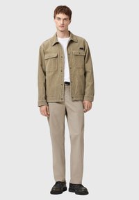Ung man i beige manchesterjacka, vit t-shirt, ljusbeige byxor, och svarta skor, som står mot en enfärgad ljusgrå bakgrund.