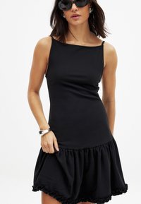 Vestido negro sin mangas con corpiño ajustado y dobladillo con volantes. Hecho de una tela suave, cuenta con finas tirantes en los hombros y una falda fluida.