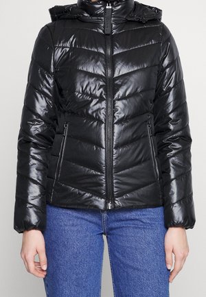 Schwarze glänzende Pufferjacke mit Chevron-Steppung und Reißverschlusstaschen, getragen über blauen Jeans, Frontansicht von Taille bis Hals.