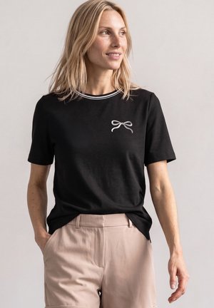 Kvinde iført en sort T-shirt med et lille hvidt sløjfedesign, parret med beige bukser, stående mod en ensfarvet lys baggrund.