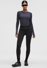 Naine seisab mustade prillide, tumedast hallist pikast varrukast topist, mustadest retuusidest, musta vöökotiga ja mustade tossudega lihtsal taustal.