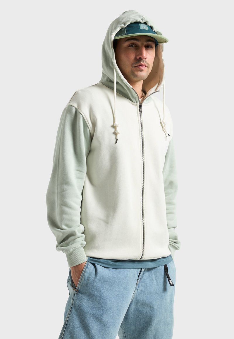 Sweat à capuche zippé avec un corps crème et des manches vert menthe ; doté de cordons de serrage et de poignets côtelés. Porté avec un pantalon en denim décontracté.