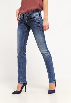 Femme portant un jean skinny bleu délavé et des chaussures à talons hauts bleu marine, debout avec une main sur la hanche devant un fond uni.