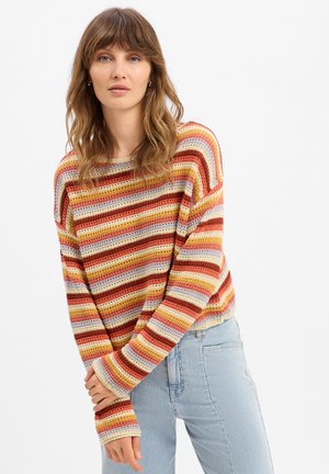 Jeune femme aux cheveux bruns mi-longs portant un pull coloré à rayures en maille et un jean bleu clair, debout devant un fond blanc.