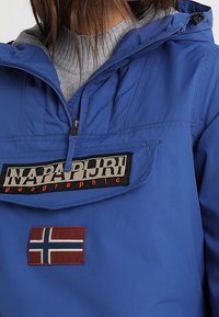 Modrá bunda Napapijri s polovičním zipem, velkou přední kapsou a norskou vlajkou na hrudi nošená přes šedý svetr.