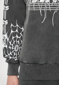 Siva majica s kapuco z belim motivom pajkove mreže na rokavu, rebrastimi manšetami in teksturo, značilno za bombažno zmes.
