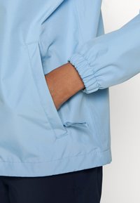 Main dans la poche d'une veste bleu clair avec poignet élastique, pantalon bleu marine visible sous l'ourlet de la veste.