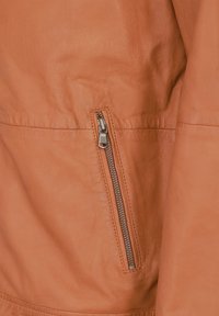 Tan læderjakke med sidelomme med sølvhardware, glat tekstur og et slankt design.