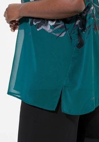 Teal gennemsigtig bluse med blomstermønster i sorte nuancer, med en sidesplit og flydende design. Båret over sorte bukser.