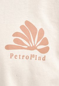Beige stof met een groot roze grafisch ontwerp dat lijkt op gestileerde bloemblaadjes, met daaronder het woord "Petrol®Ind" in een eenvoudige lettertype.