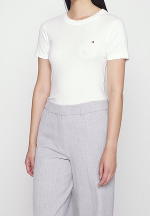 Kobieta ubrana w dopasowany biały t-shirt z krótkim rękawem i jasnoszare, wysokie taliowane spodnie na białym tle.