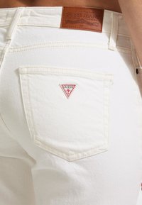 Vaqueros de mezclilla blancos con un logo triangular rojo de "GUESS?" en el bolsillo trasero y una etiqueta de marca de cuero marrón en la cinturilla.