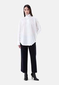 Camicia bianca a maniche lunghe con colletto alto, abbinata a pantaloni blu scuro su misura e stivaletti neri con tacco, su uno sfondo neutro.